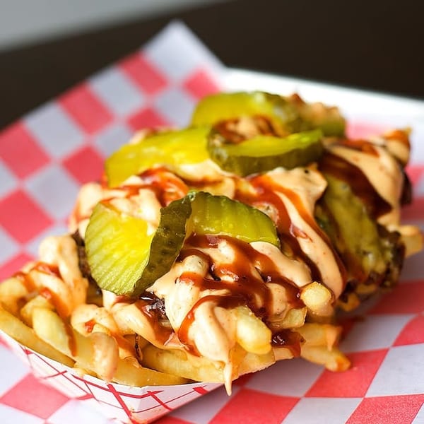 Loaded fries with cheese sauce, BBQ sauce, and pickles in a red and white checkered paper tray.