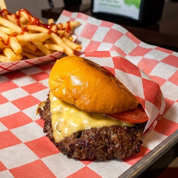 A cheeseburger with tomato and a side of ketchup-drizzled fries on red and white checkered paper.
