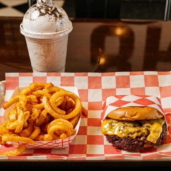 A cheeseburger, curly fries, and an Oreo milkshake on a metal tray with red and white checkered paper.